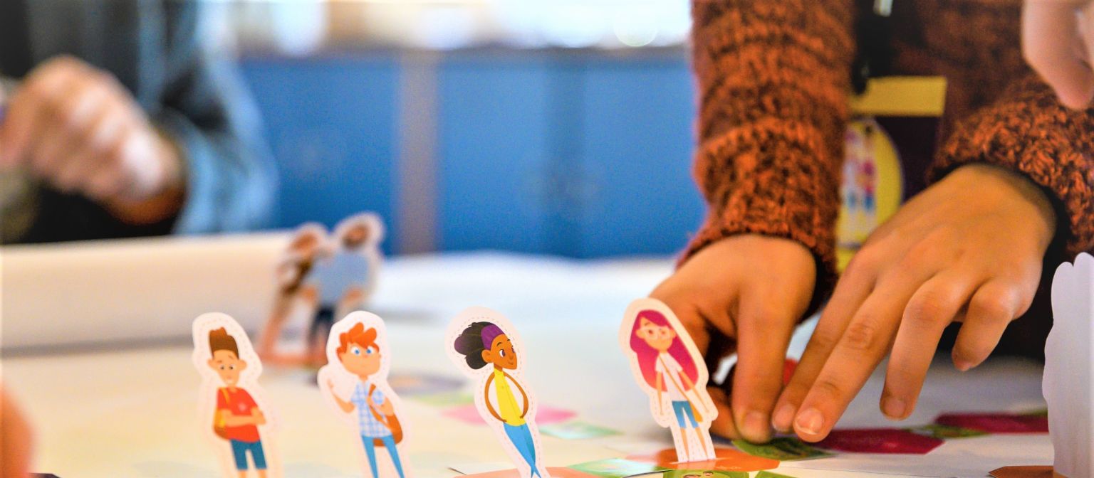 Ein Foto zeigt Hände, die Figuren von Schüler*innen auf einem Spielfeld bewegen. Darunter steht der Text: Finanzbildung: Wer früh den richtigen Umgang mit Geld erlernt, ist später besser vor Überschuldung geschützt.