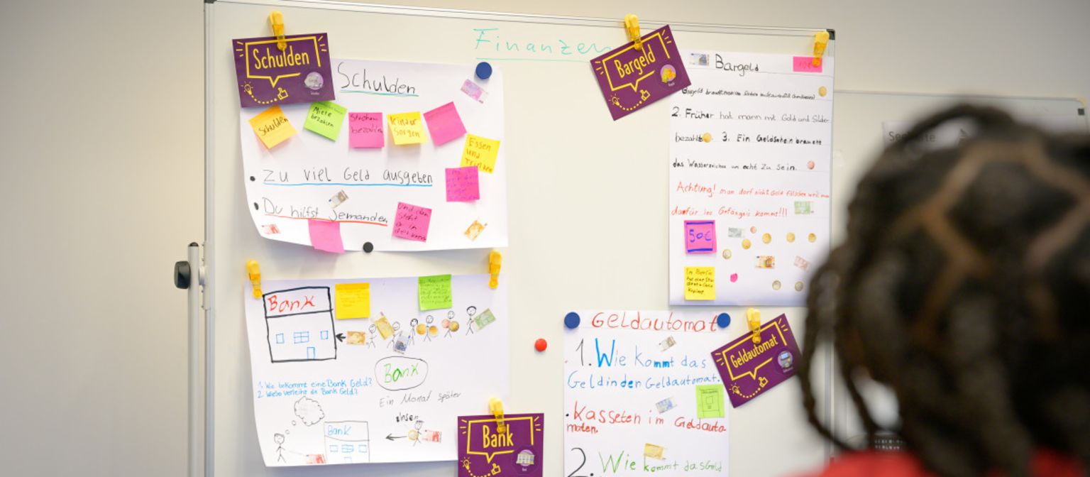 Ein Foto zeigt Post-its und Zettel mit Notizen an einer Tafel, auf denen Begriffe wie „Schulden“, „Bargeld“, „Bank“ und „Geldautomat“ stehen. Darunter steht der Text: „Im Programm ManoMoneta der EOS eigenen finlit foundation können Schüler im Alter von 9 bis 13 Jahren Wissen über Finanzprodukte erwerben.“