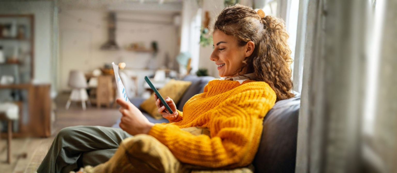 Une jeune femme souriante aux cheveux bouclés, vêtue d'un pull jaune confortable, est assise sur un canapé dans son salon et regarde son smartphone tout en tenant un document papier.