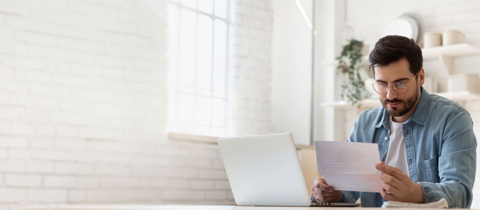 Mann mit Brille arbeitet am Laptop und prüft Schuldenregulierungsdokument in hellem Zuhause