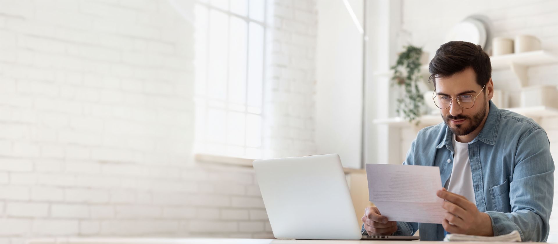Mann mit Brille arbeitet am Laptop und prüft Schuldenregulierungsdokument in hellem Zuhause