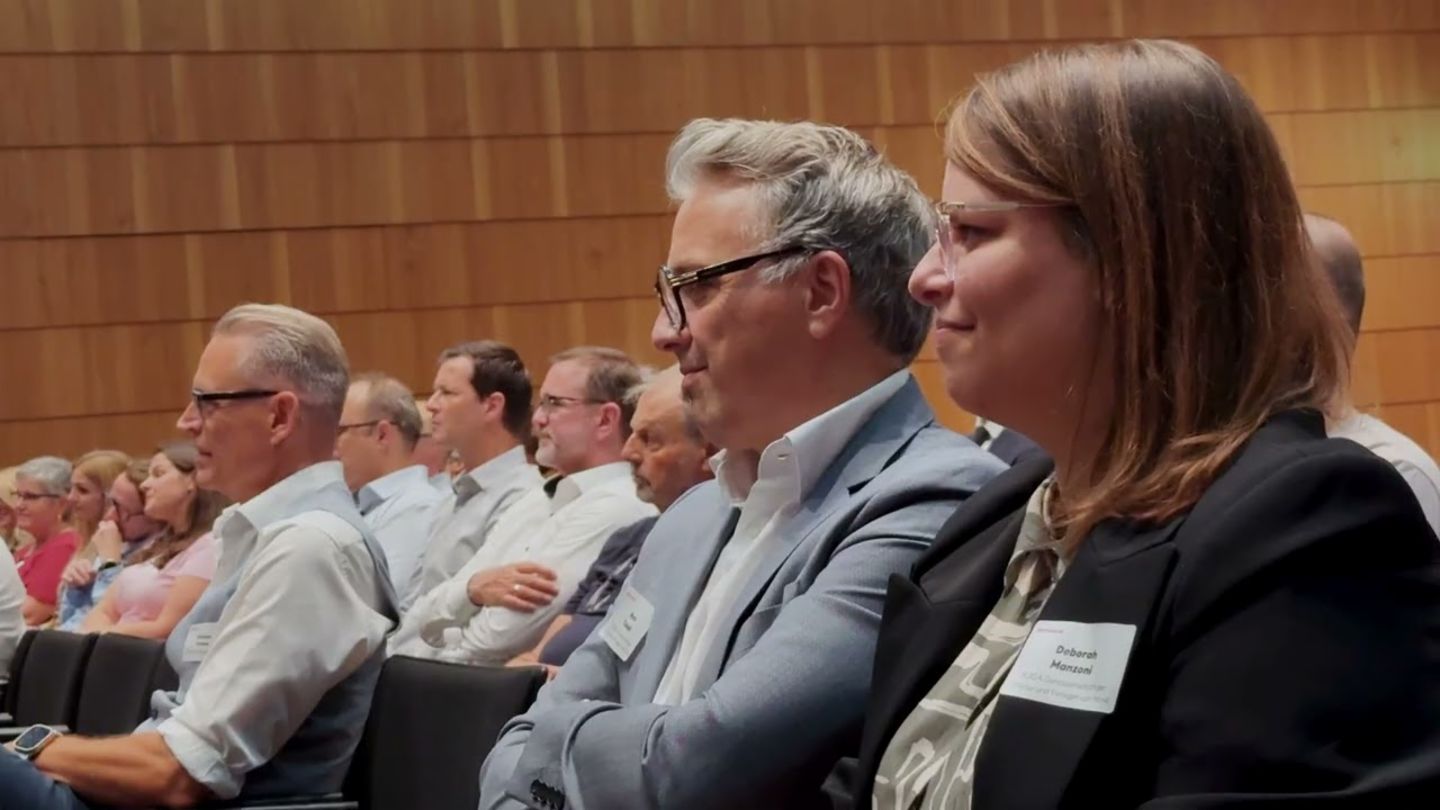 Publikum im Konferenzsaal, im Fokus zwei aufmerksam zuhörende Gäste mit Namensschild vor einer Holzwand, im Hintergrund weitere Zuschauer.