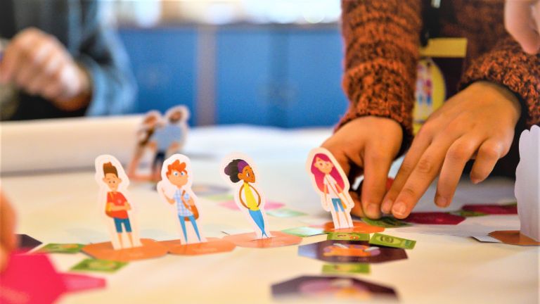 Ein Foto zeigt Hände, die Figuren von Schüler*innen auf einem Spielfeld bewegen. Darunter steht der Text: Finanzbildung: Wer früh den richtigen Umgang mit Geld erlernt, ist später besser vor Überschuldung geschützt.