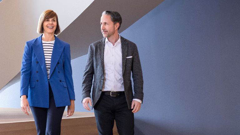 Le PDG en blazer gris et le CFO du Groupe EOS en blazer bleu traversent un bureau moderne. L'espace dispose de grandes fenêtres, d'un mur courbé et de lampes suspendues au design élégant.