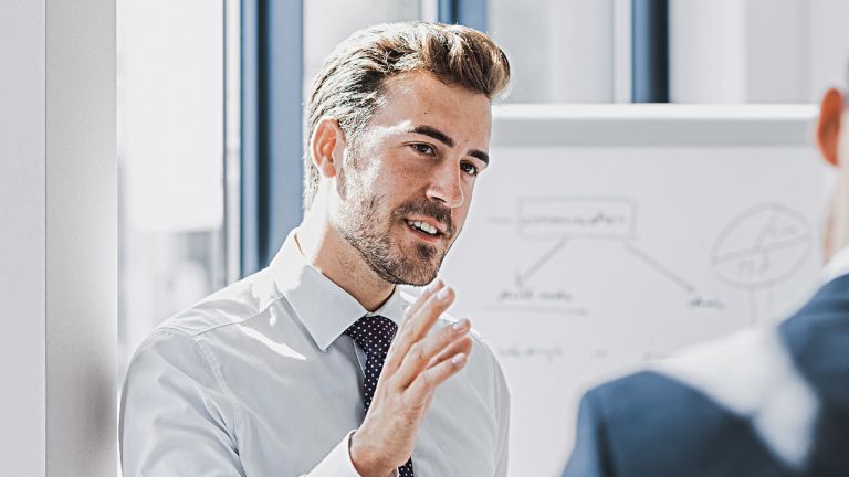 On voit un homme en tenue professionnelle expliquer quelque chose avec une expression amicale et engagée. Face à lui se tient une autre personne qui l’écoute attentivement. On aperçoit en arrière-plan un tableau blanc avec un croquis.