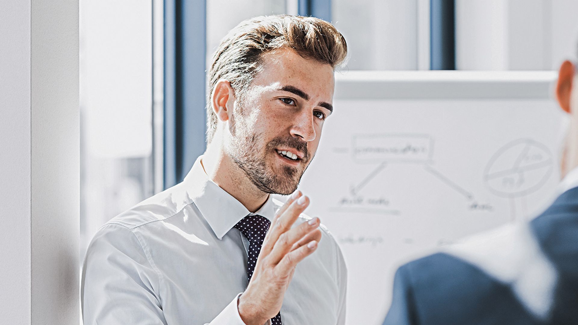 Zu sehen ist ein Mann in Business-Bekleidung, der mit freundlich-engagiertem Ausdruck etwas erklärt. Vor ihm steht eine andere Person, die offensichtlich Zuhörer ist. Im Hintergrund ist ein White-Board mit einer Skizze zu sehen.