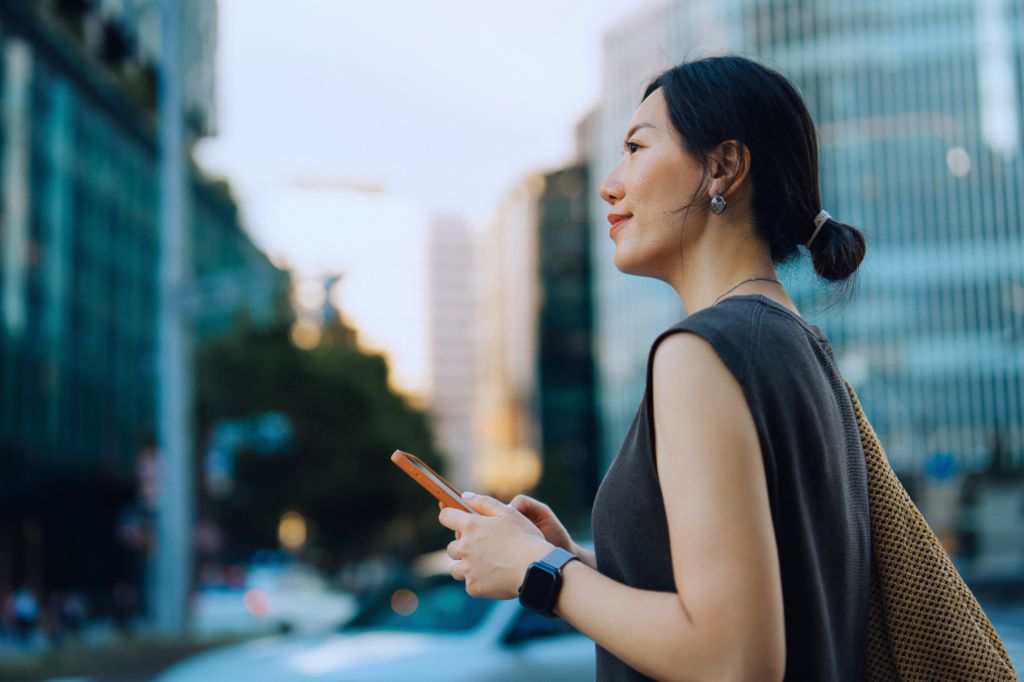 Eine junge Frau steht draußen in einer urbanen Umgebung, hält ihr Smartphone und blickt lächelnd nach vorne. Im Hintergrund sind moderne Gebäude und Bäume zu sehen.