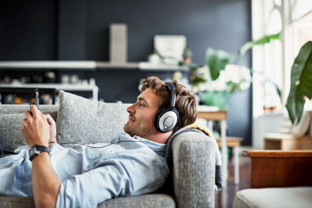 Ein junger Mann liegt auf einer Couch in seinem Wohnzimmer, trägt Kopfhörer und schaut lächelnd auf sein Smartphone, während er Musik oder einen Podcast genießt.