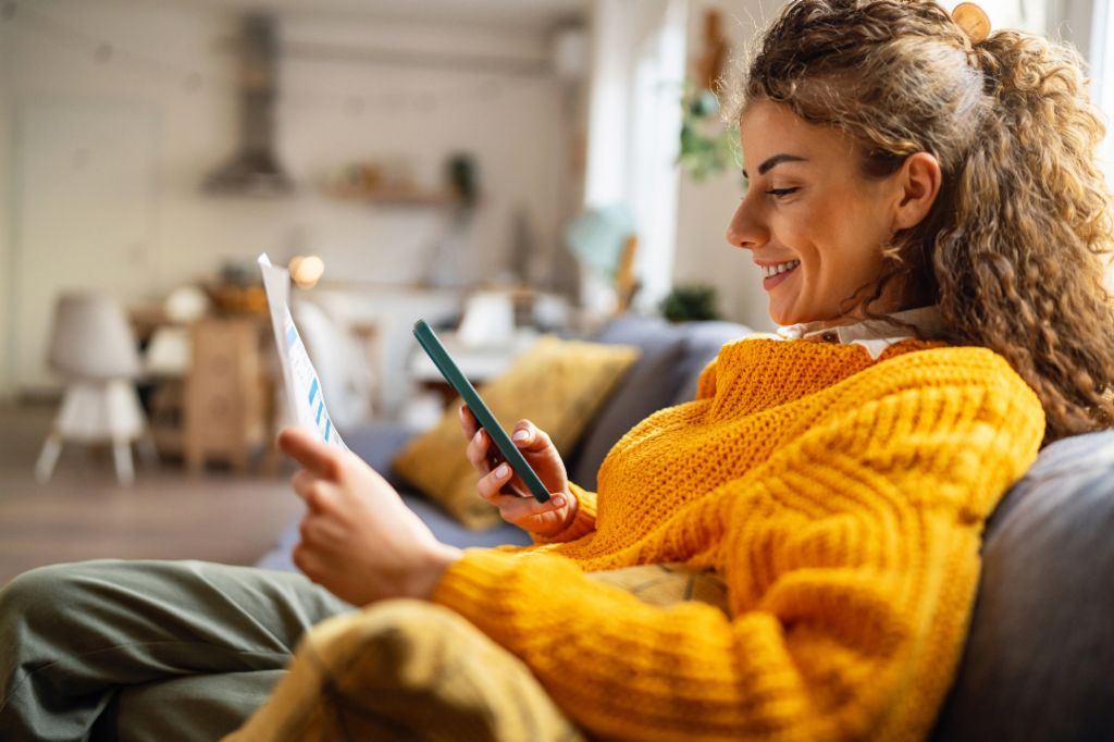 Eine lächelnde junge Frau mit lockigen Haaren in einem gemütlichen gelben Pullover sitzt auf dem Sofa in ihrem Wohnzimmer und schaut auf ihr Smartphone, während sie ein Papierdokument in der Hand hält.