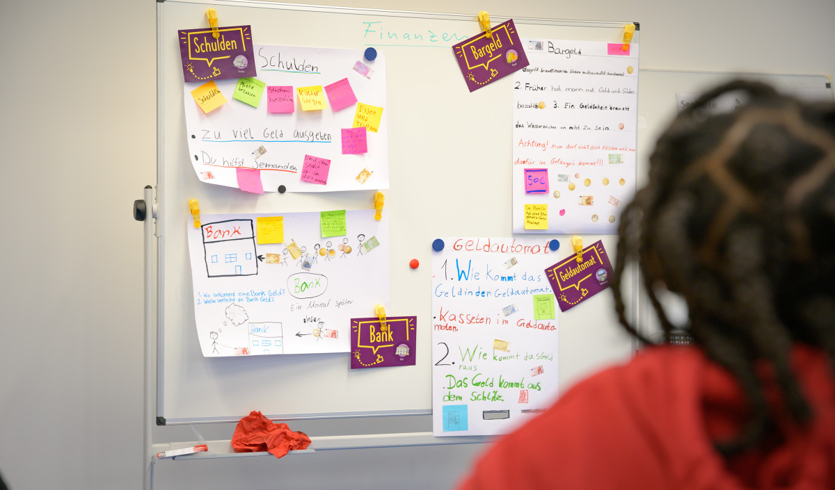 Ein Foto zeigt Post-its und Zettel mit Notizen an einer Tafel, auf denen Begriffe wie „Schulden“, „Bargeld“, „Bank“ und „Geldautomat“ stehen. Darunter steht der Text: „Im Programm ManoMoneta der EOS eigenen finlit foundation können Schüler im Alter von 9 bis 13 Jahren Wissen über Finanzprodukte erwerben.“