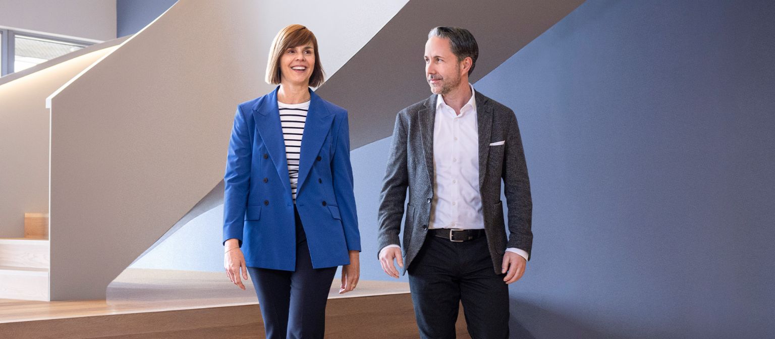 Der CEO im grauen Blazer und die CFO der EOS Gruppe in blauem Blazer gehen in einem modernen Büro. Der Raum hat große Fenster, eine geschwungene Wand und stilvolle hängende Lampen.