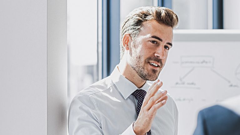 Zu sehen ist ein Mann in Business-Bekleidung, der mit freundlich-engagiertem Ausdruck etwas erklärt. Vor ihm steht eine andere Person, die offensichtlich Zuhörer ist. Im Hintergrund ist ein White-Board mit einer Skizze zu sehen.