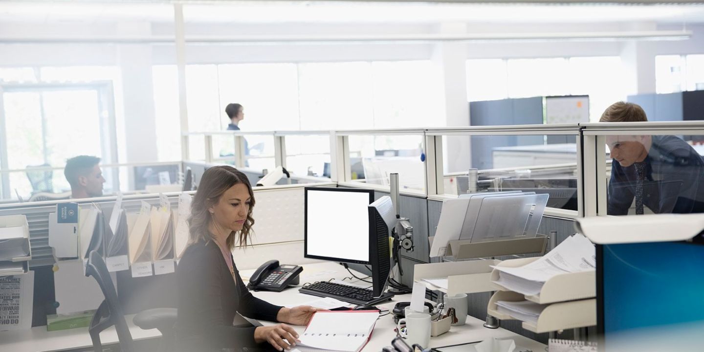 Digitalisierung: Drei Mitarbeiter sind in einem Großraumbüro, eine Frau sitzt zwischen Papieren und ihrem Computer