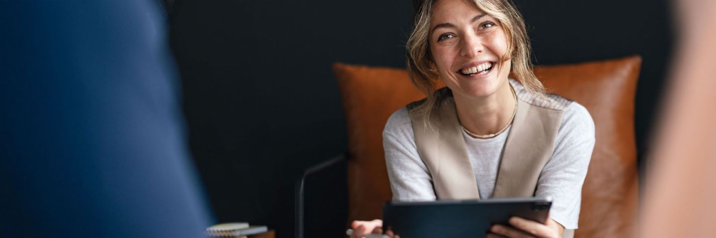 Image of a young woman laughing openly and holding a tablet computer. She is seated on a leather armchair in front of a dark backdrop.
