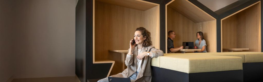 Lächelnde Frau sitzt in einer modernen Büro-Lounge, telefoniert und arbeitet am Laptop. Im Hintergrund arbeiten zwei weitere Personen.
