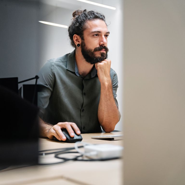 Un homme barbu avec les cheveux longs attachés en chignon est assis devant un ordinateur, complètement absorbé par son travail.
