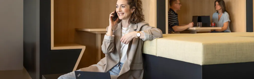 Une personne souriante est assise dans un salon de bureau moderne, parle au téléphone et travaille sur un ordinateur portable. En arrière-plan, deux autres personnes travaillent.