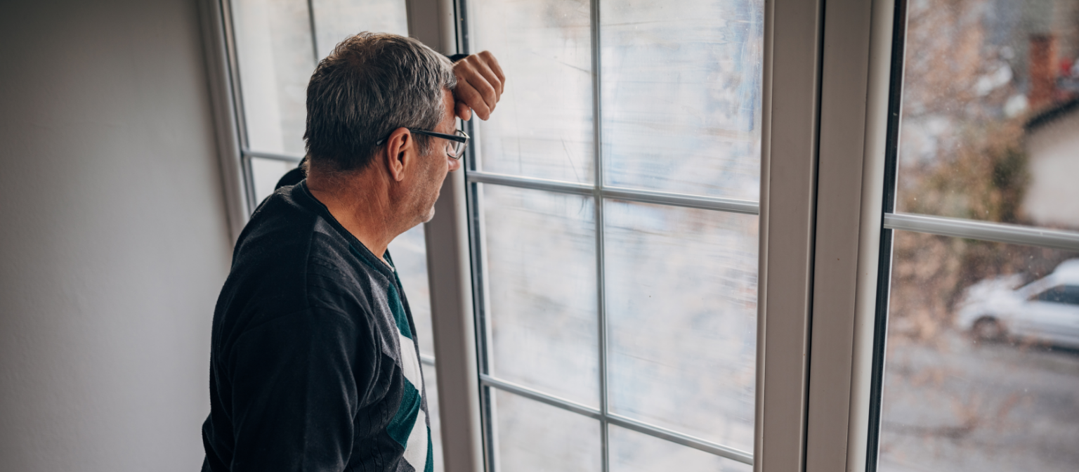 Recouvrement équitable des créances : un débiteur d'âge moyen vêtu d'une veste olive regarde par la fenêtre avec inquiétude.