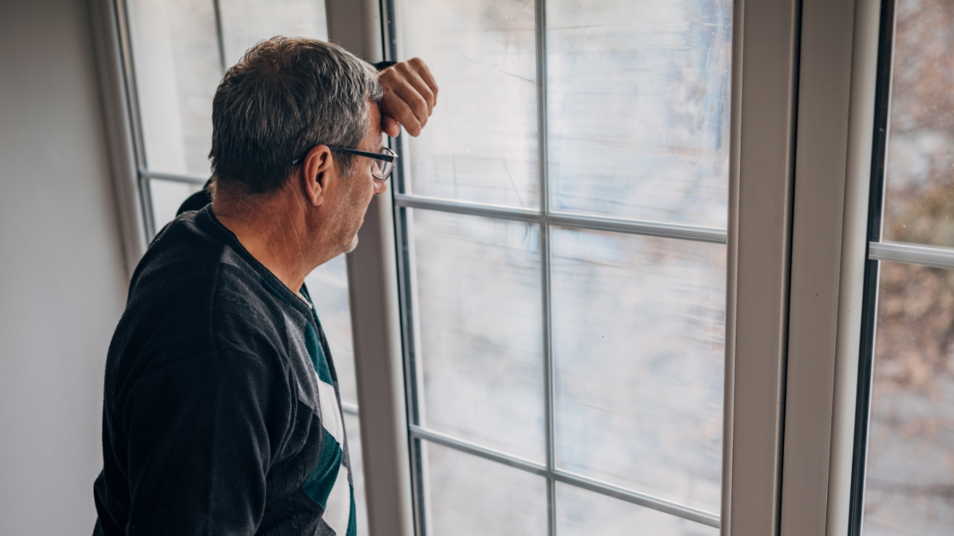 Recouvrement équitable des créances : un débiteur d'âge moyen vêtu d'une veste olive regarde par la fenêtre avec inquiétude.