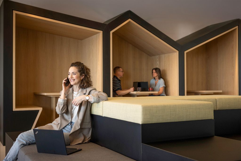 Une personne souriante est assise dans un salon de bureau moderne, parle au téléphone et travaille sur un ordinateur portable. En arrière-plan, deux autres personnes travaillent.