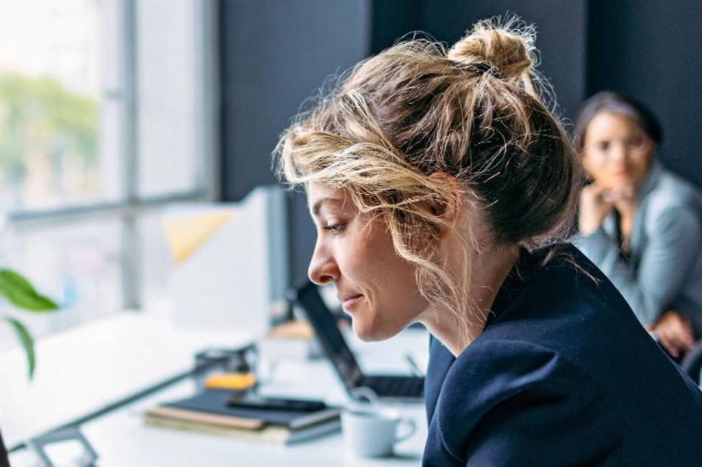 L'image montre une femme assise à un bureau dans un bureau moderne, portant un blazer sombre tout en se concentrant sur son ordinateur portable, avec une plante et quelques fournitures de bureau visibles sur la table, et une autre personne assise en arrière-plan.